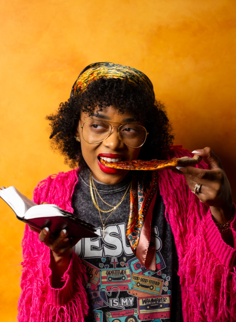 Image of Shanika standing in front of an orange wall eating a slice of pizza with the bible in another hand.