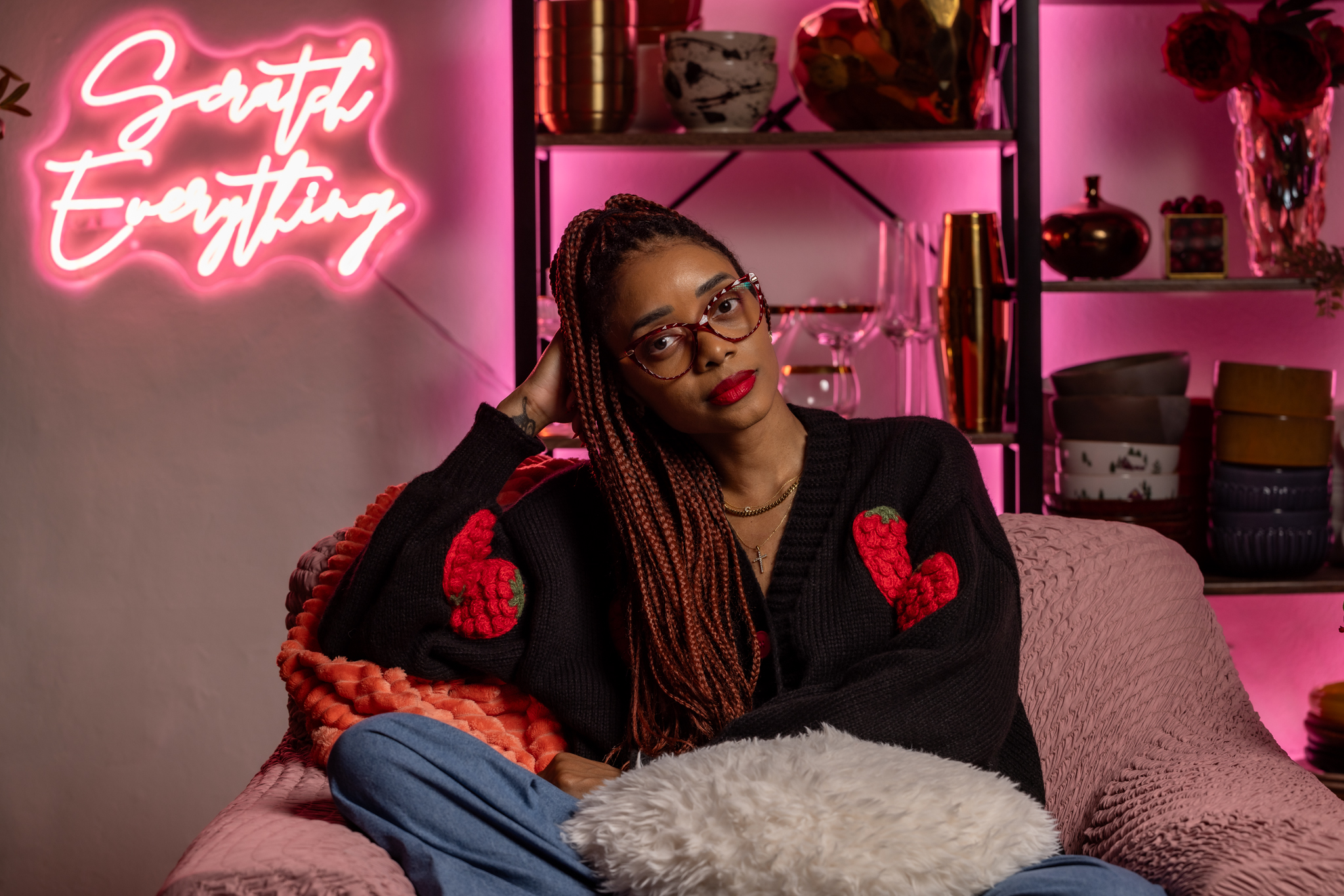 An image os Shanika sitting on a couch with a red/black sweater and pink background.