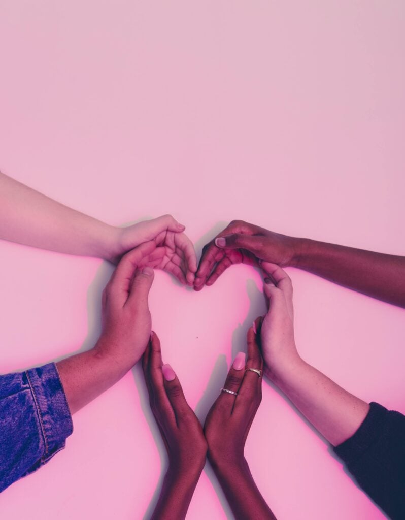 Different hands forming in a shape of a heart on a pink background.