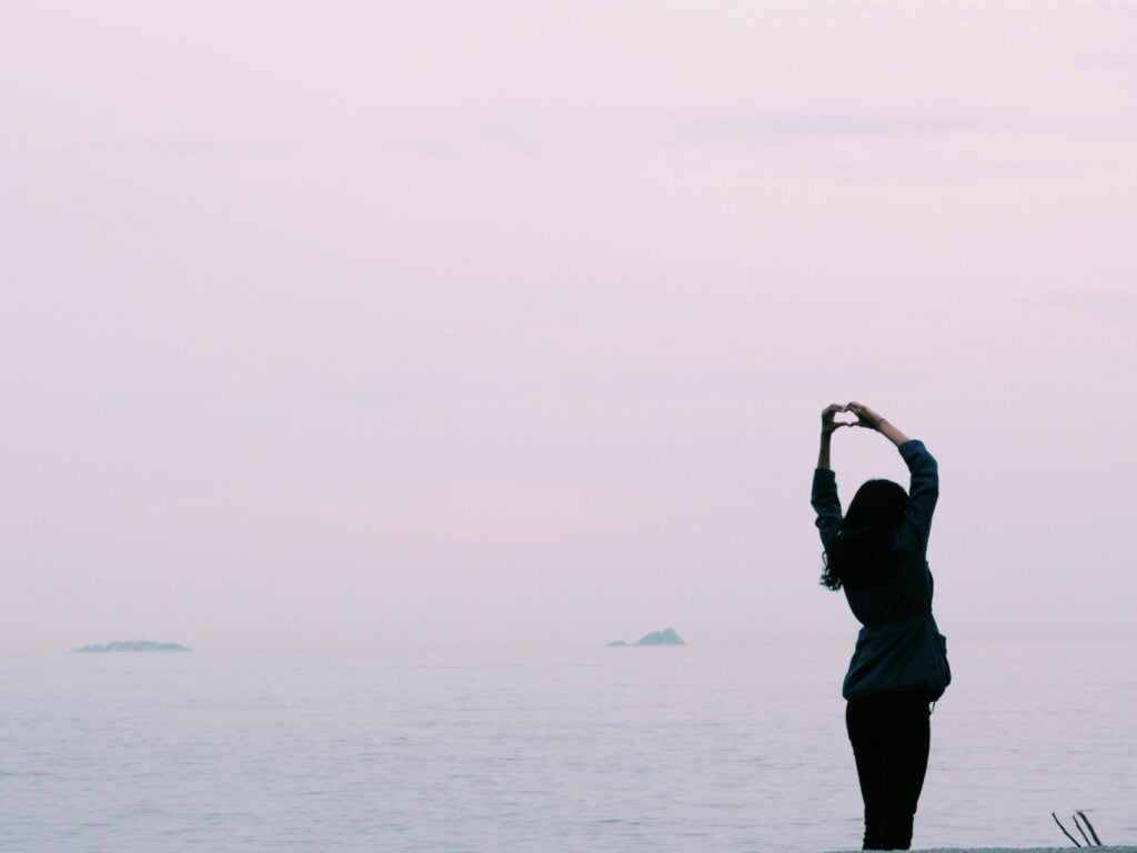 A woman standing in front of water with hands raised and in the shape of a heart.