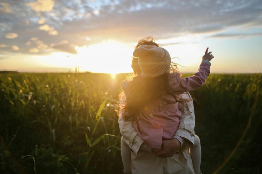 A mother holding her daughter in hand in a field with a sunset.