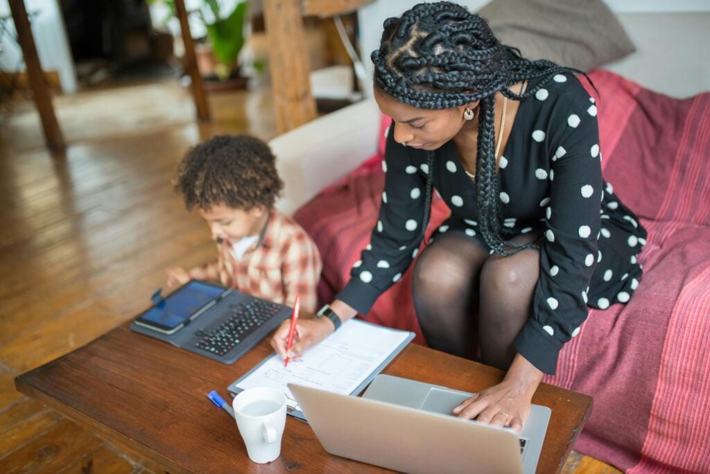 A mother sitting and working on computer with son next to her on tablet.
