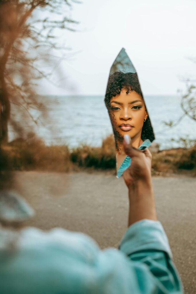 A woman holding a piece of glass and looking into broken mirror.