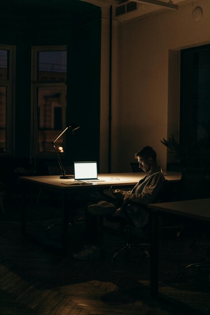 Someone sitting in the dark with a laptop open and a lamp.