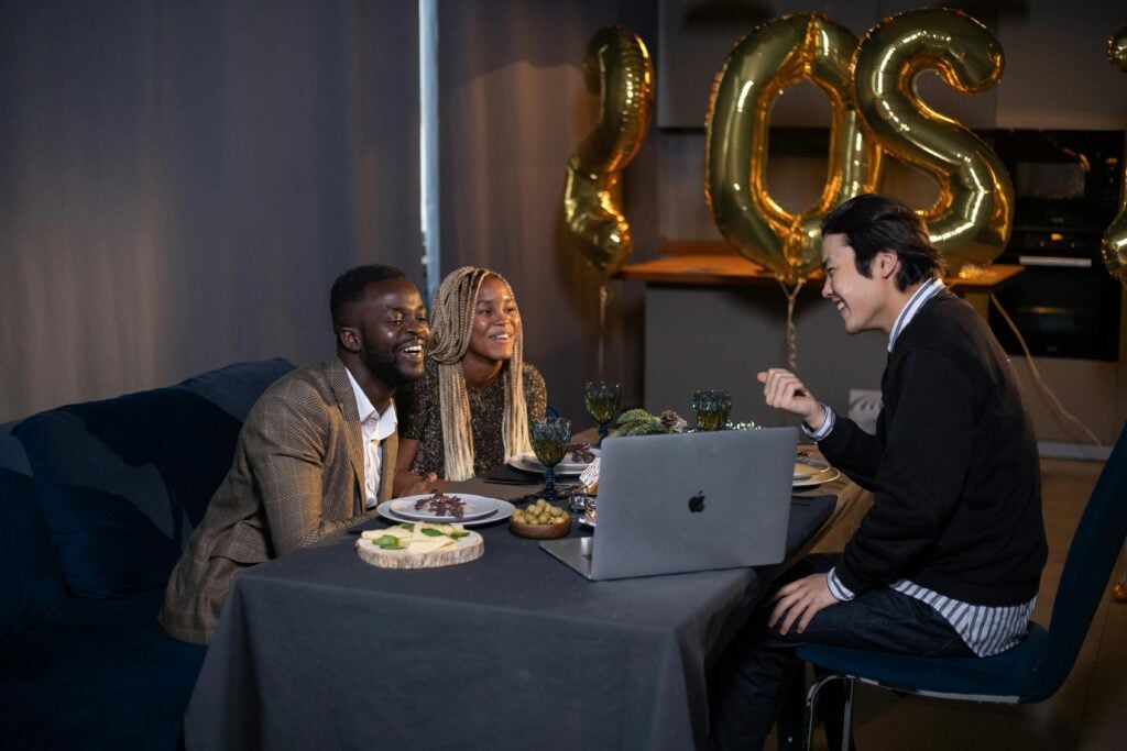 Three people at a table laughing with balloons and a laptop.