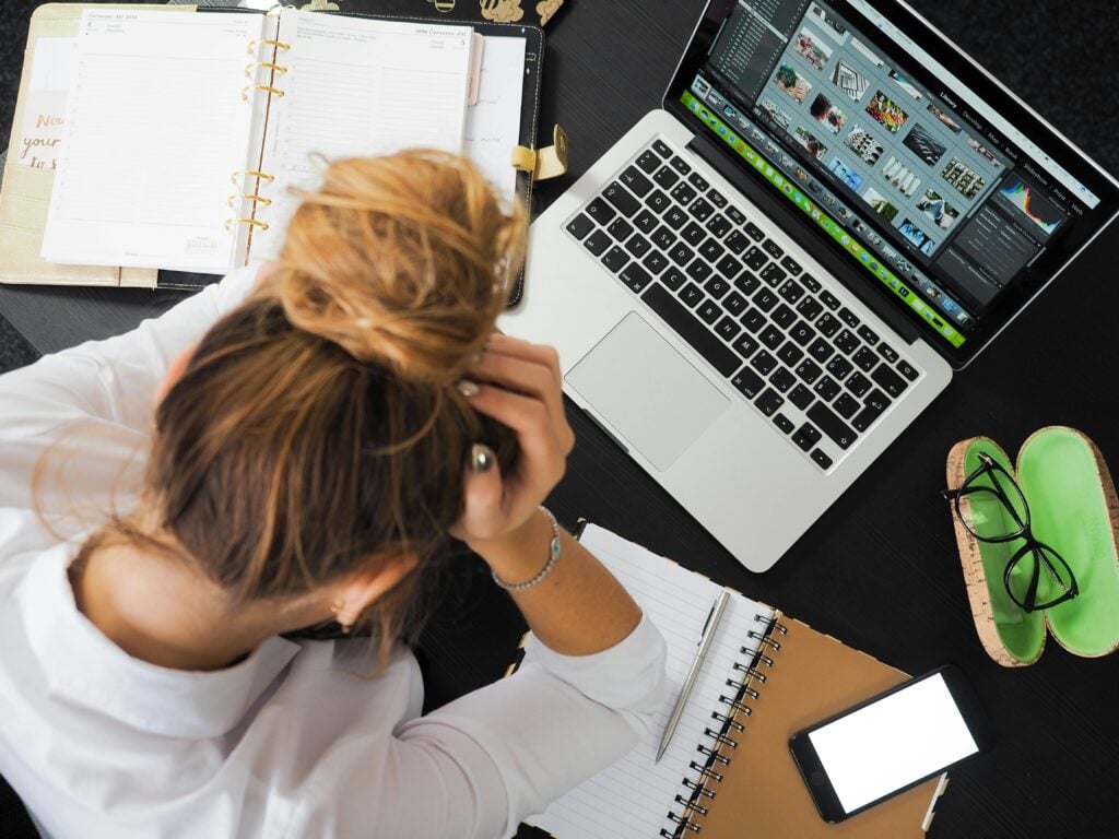 A woman with her head in hands next to a laptop and full desk.