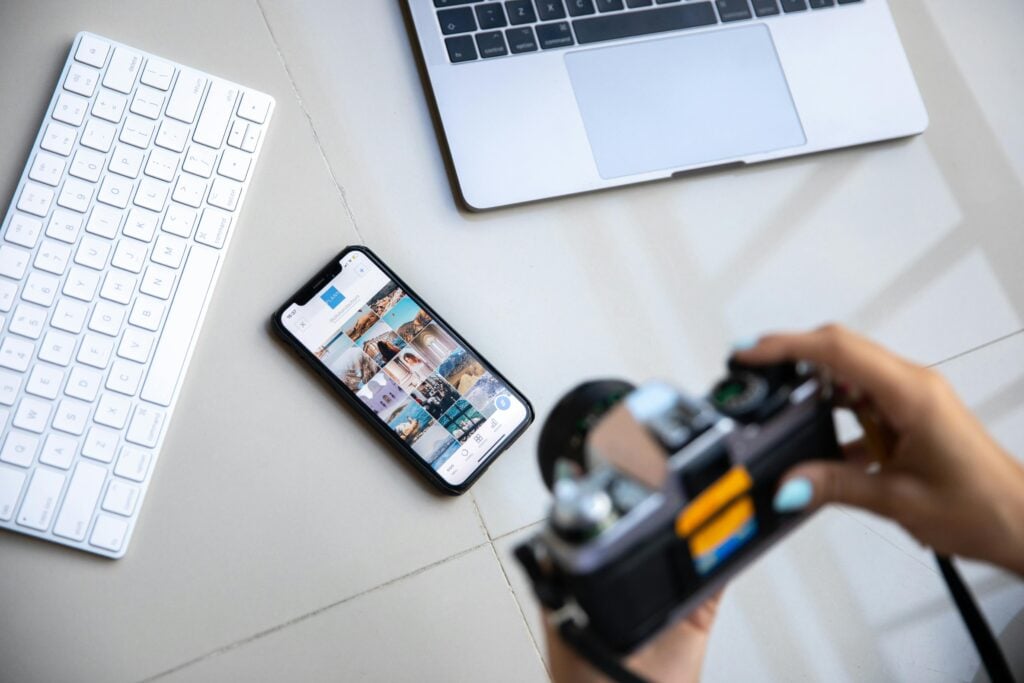 Woman hands holding a camera in front of a table with a smart phone, keyword, and laptop.