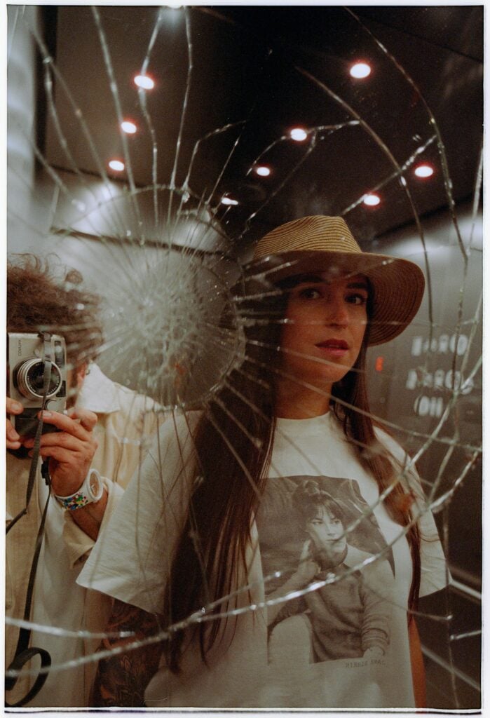 A female looking in a broken mirror while a guy behind her takes photos with a camera.