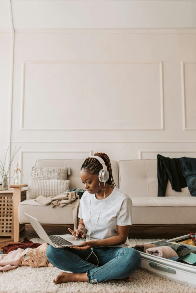 A woman sitting on the floor with headphones and a laptop and things around her.
