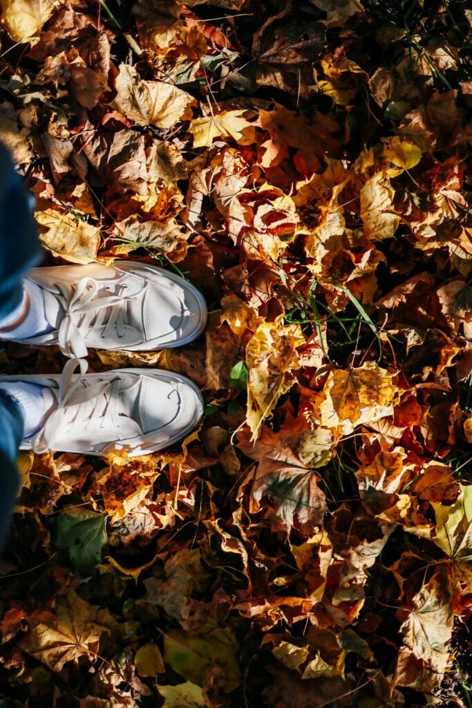 Fall leaves on the ground with someone's feet standing with white sneakers.