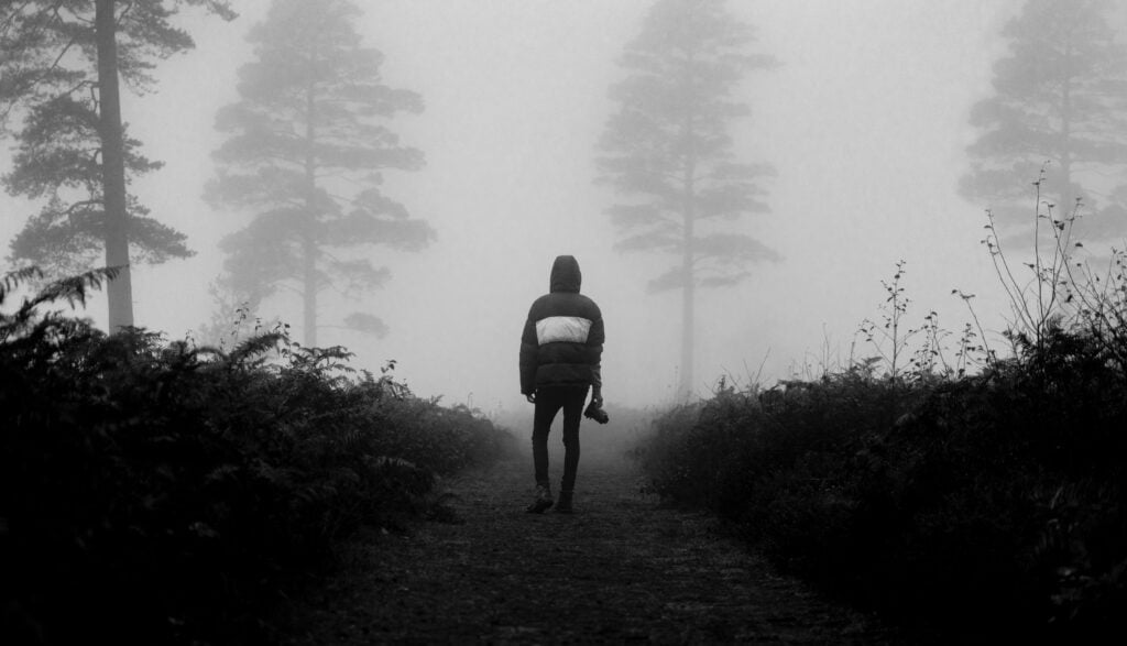 A guy walking down a path in a woody area filled with mist and holding a camera in hand.