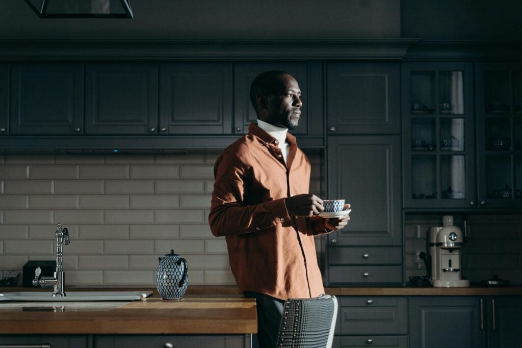 A man standing in a kitchen with a brown jacket and holding a cup of tea or coffee.