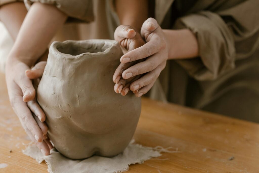 Two set of hands molding together clay in shape of a jar.