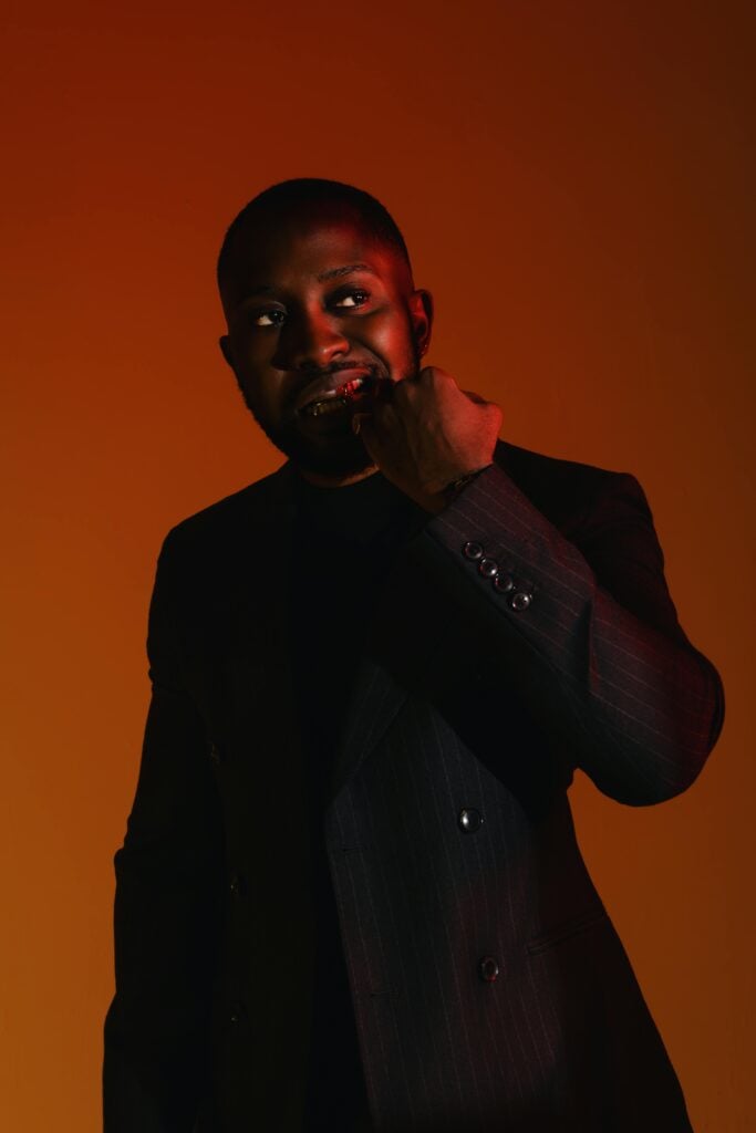 A man in a black suit holding mouth in front of a dark red backgorund.