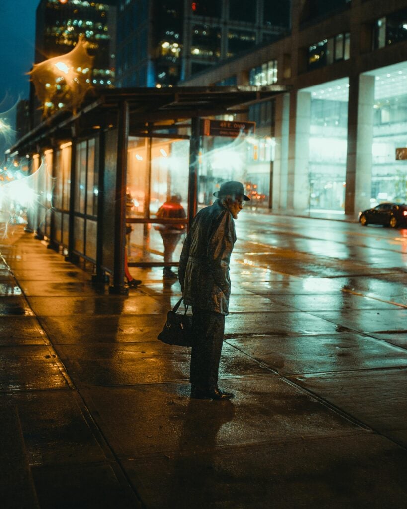 An old man standing on the street at a bus stop in the night.
