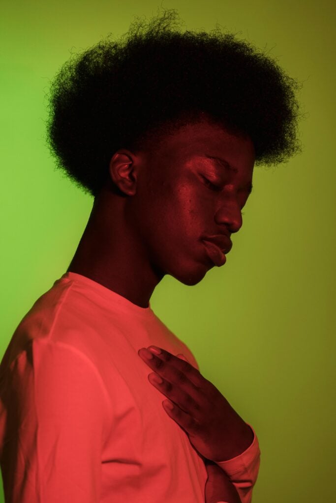 A young man in a neon shirt closing eyes with hand on chest in front of lime colored wall.