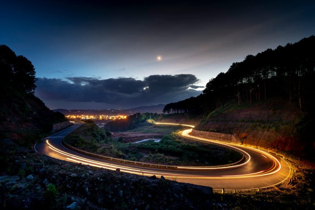 A curved road in the midst of mountains and a horizon with lights at night.