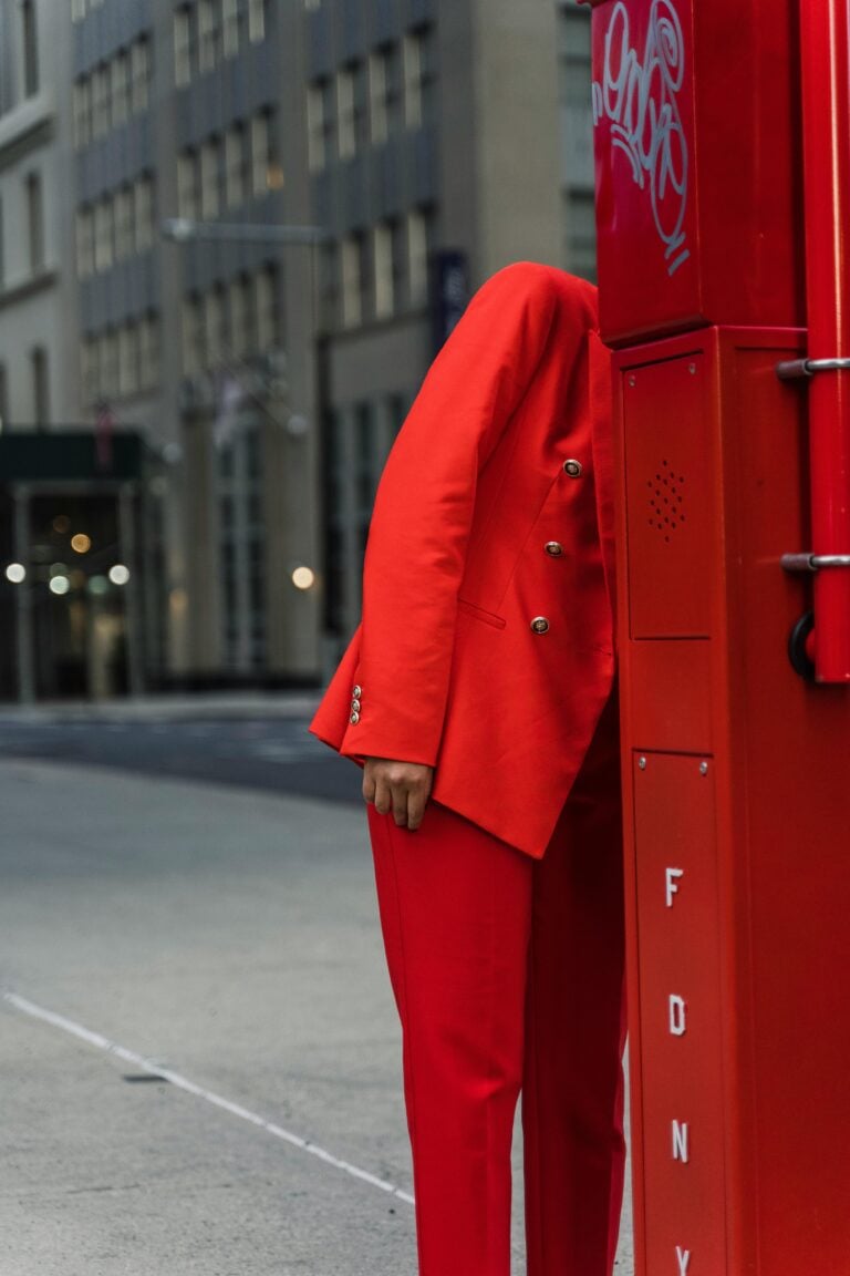 Someone in a red suit hiding their face behind a red post outside a city.