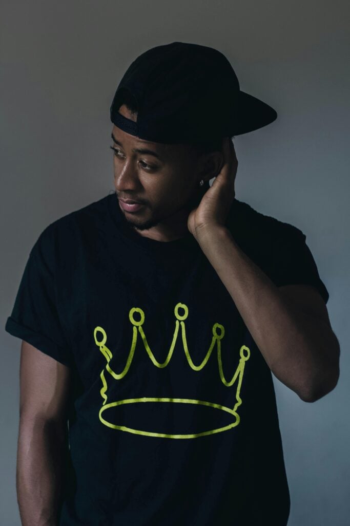 A guy looking to the side wearing a black shirt with a crown on it and a cap.