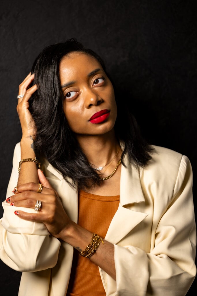 Shanika looking to the side slightly with hand holding head with a beige blazer and brown shirt.