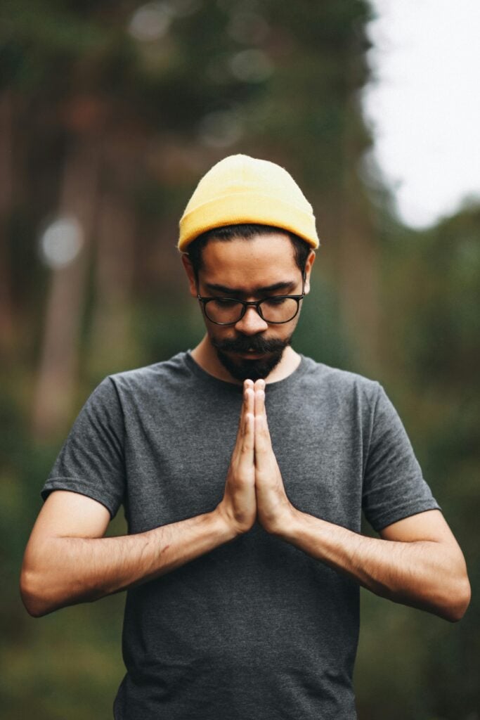 A guy with glasses, beanie, and t-shirt looking down with hands closed in prayer.