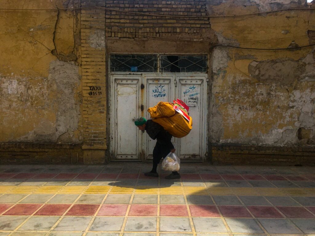 A man walking with a heavy loaded bag on his back in front of a brick building.