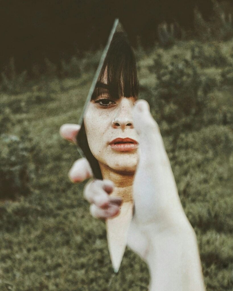 A woman holding a piece of mirror looking at her reflection. 