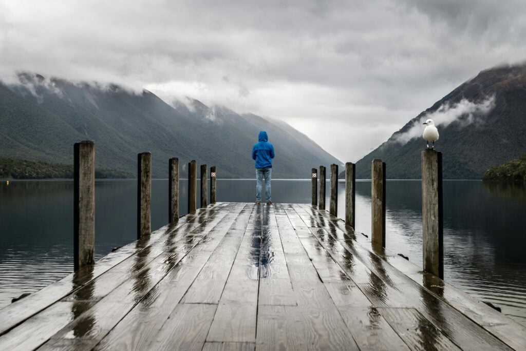 A guy standing at the end of a dock looking at the water and mountain view.
