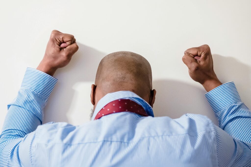 A guy in a shirt and tie facing the wall with both fists in the air.