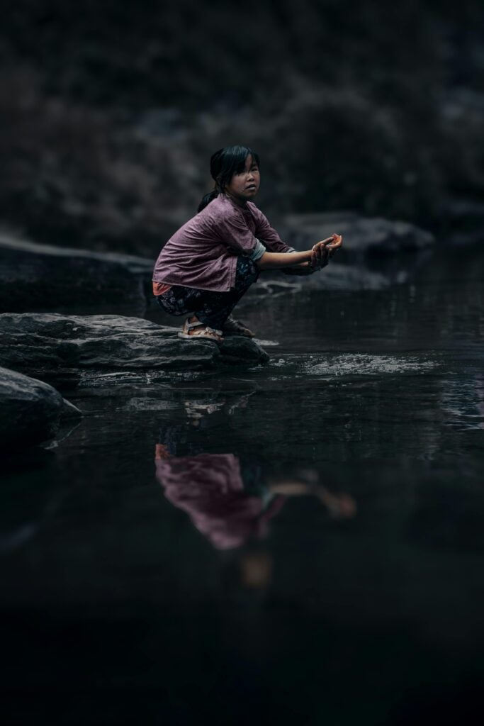 A child bending at the edge of a rock near water.