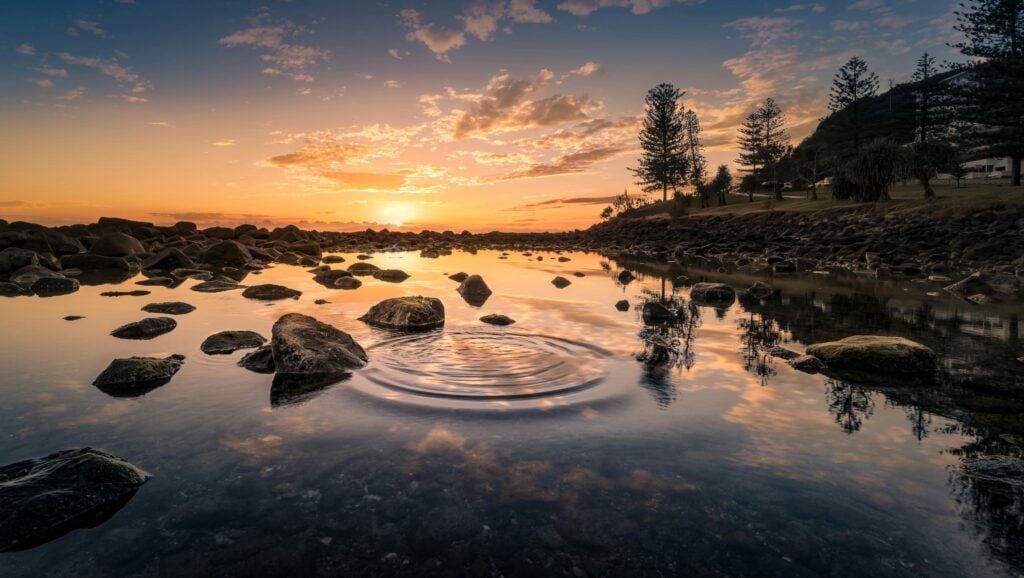 Water with a view of sunset and rocks with greenery.