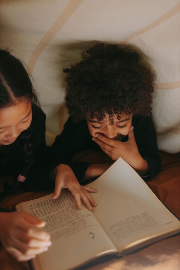Two kids reading a book together under sheets.