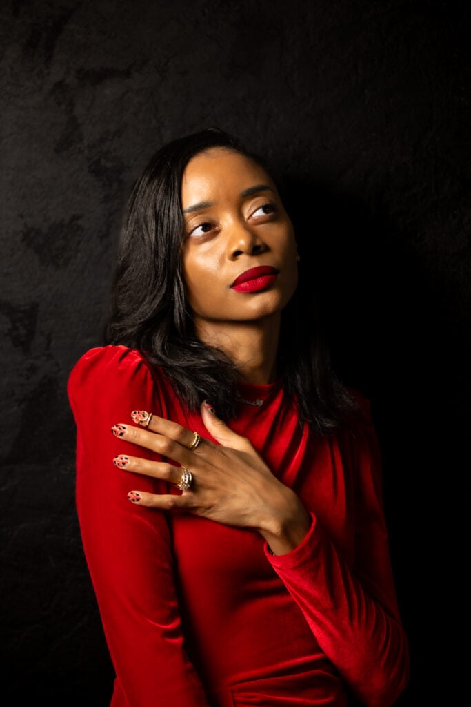 An image of Shanika turned slightly to the side in a red dress looking up in front of a black background.