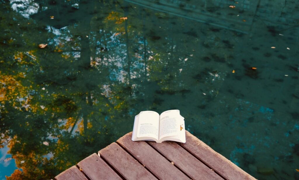 An open book on a deck in front of water.