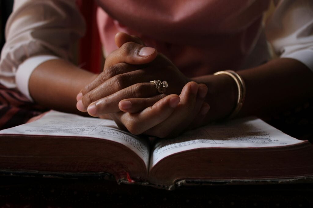 A woman's hands folded atop an opened bible in prayer position.