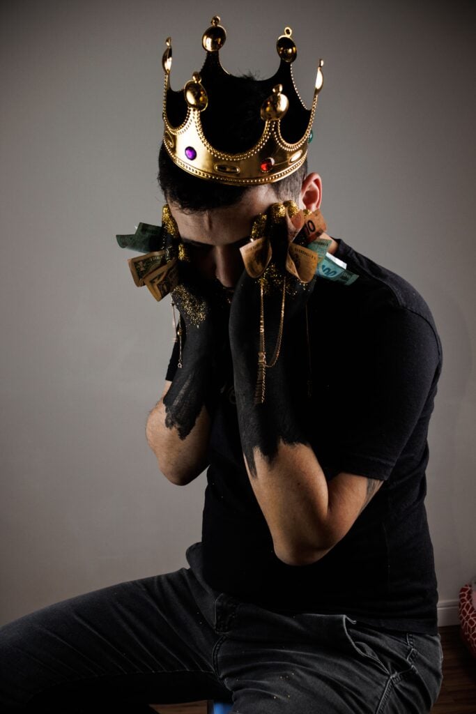 A man with a black shirt sitting with a crown on his head and face in his hands.